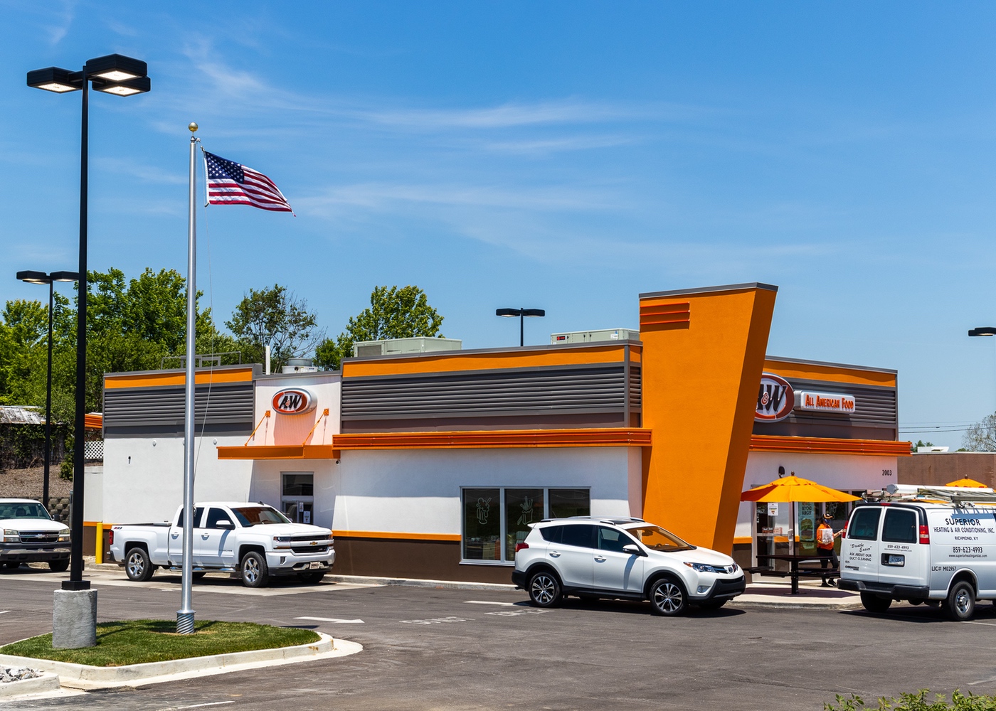 Exterior side view photo of A&amp;amp;W Restaurant showing cars in drive-thru and team member walking bag out to car.